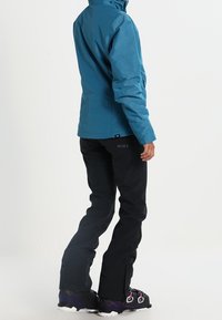 Türkisfarbene wasserdichte Jacke mit tailliertem Design, getragen über schwarzen Skihosen und Stiefeln. Die Jacke hat einen hohen Kragen und ein Logodetail am Bund.