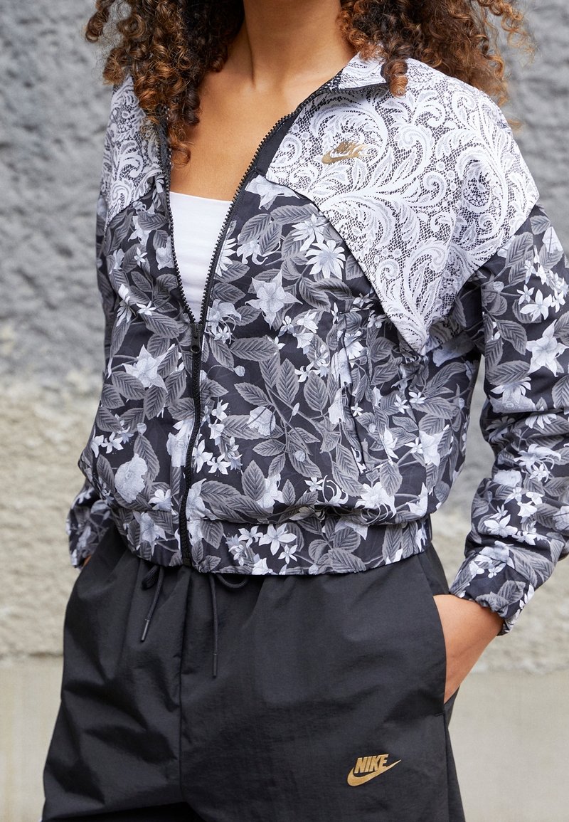 Bomberjacka med blommönster som har svarta och vita designer, dragkedjestängning och elastiska muddar. Nike-logotyp i guld på nedersta vänstra sidan.