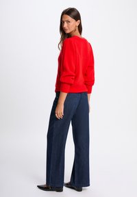 Roter, strukturierter Strickpullover mit überschnittenen Schultern; kombiniert mit hoch taillierten, weit geschnittenen Jeans aus dunklem Denim und spitzen schwarzen Ballerinas.