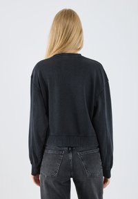 Felpa nera corta con polsini e orlo a costine, caratterizzata da una texture morbida. Abbinata a jeans grigio scuro che mostrano dettagli delle tasche posteriori.