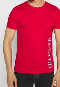 Červené bavlněné tričko s kulatým výstřihem a krátkými rukávy. Na levé straně je vertikálně potištěný text 'HILFIGER' bílou barvou.