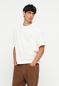 White cotton t-shirt with a relaxed fit, featuring a small peach-colored logo on the chest, paired with brown corduroy pants.