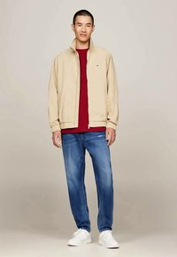 Beige lightweight jacket, red t-shirt, blue jeans, and white sneakers. Jacket has elastic cuffs and hem; denim shows a faded wash.