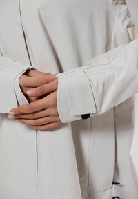 Vit långärmad plagg med breda manschetter. Tyget verkar slätt och något glänsande, med subtil sömdetaljering och slits.