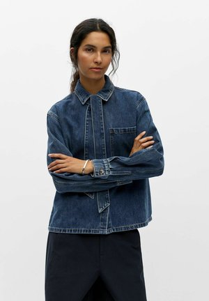 Femme aux cheveux foncés portant une veste en jean bleue et un pantalon noir, debout les bras croisés devant un fond clair uni.
