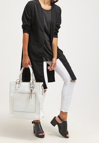 Cardigan lungo nero, canotta nera, jeans bianchi strappati, scarpe con tacco a blocco aperte nere e borsa in pelle bianca con rifiniture argentate.