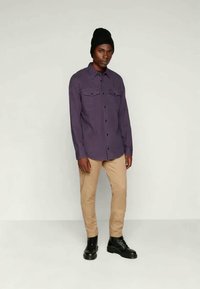 Camicia a maniche lunghe viola con bottoni e due tasche sul petto, abbinata a pantaloni beige e stivali neri stringati. Indossata con un berretto nero.