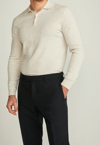 Helles beige Langarm-Polohemd mit geradem Kragen und Knopfverschluss, getragen zu dunklen Anzughosen, mit einer glatten Textur.