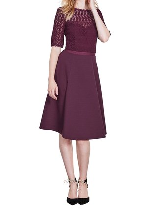 Vestido burdeos con corsé de encaje, mangas cortas y una cintura ceñida que fluye hacia una falda a la altura de la rodilla; combinado con tacones negros.