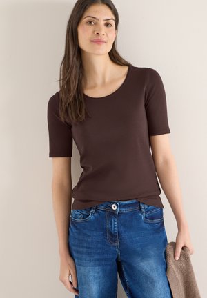 Young woman with long dark hair wearing a dark brown short-sleeve top and blue jeans, holding a brown knit sweater in one hand.