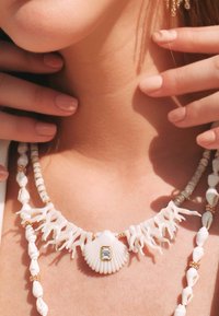 Collier blanc mettant en avant un pendentif en coquillage central avec une gemme rectangulaire, accentué par des branches rappelant le corail et de petites perles.