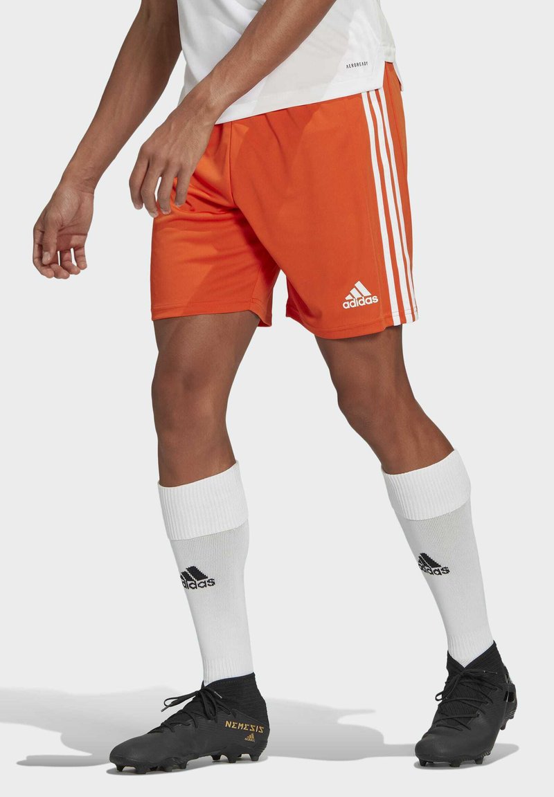 Orangene Sportshorts mit weißen Seitenstreifen, kombiniert mit weißen knielangen Socken mit einem schwarzen Logo und schwarzen Fußballschuhen.