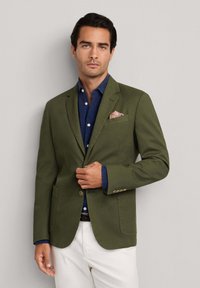 Mann, der einen Knopf an einem olivgrünen Blazer über einem marineblauen Hemd und weißen Hosen vor hellem einfarbigem Hintergrund anpasst.