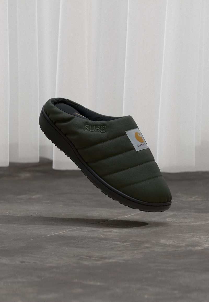 Grøn quiltet hjemmesko med en sort sål. Har et logo-patch og subtil branding. Slank, lavprofil design, lavet af blødt stof.