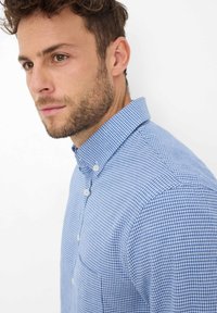 Man met lichte baard in een blauw-wit geruit overhemd, links kijkend tegen een vlakke witte achtergrond.