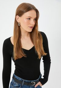 Schwarz geripptes Langarmoberteil mit V-Ausschnitt, kombiniert mit hochtaillierten blauen Jeans und einem schwarzen Gürtel mit einer silbernen Schnalle.