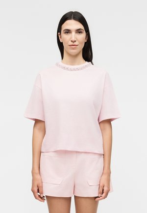 Γυναίκα με καστανά ίσια μαλλιά φοράει ροζ, διακοσμημένη μπλούζα με κοντά μανίκια και ταιριαστό σορτς, στέκεται μπροστά σε ένα απλό φόντο.