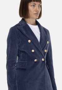 Blazer doppiopetto in velluto blu navy con bottoni dorati, ampie revers, tasche laterali, vestibilità strutturata e texture liscia.