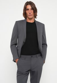Gray tailored suit with a single-button jacket and slanted pockets, paired with a black crew-neck t-shirt. Smooth fabric, fitted silhouette.