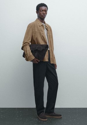 Young man wearing tan suede jacket, gray shirt, black pants, and brown shoes, holding a large dark brown folded bag, standing against plain background.