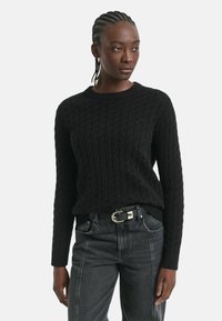Junge Person mit geflochtenen Haaren, die einen schwarzen Kabelstrickpullover und dunkelgraue Jeans mit einem schwarzen Gürtel trägt, steht vor einem weißen Hintergrund.
