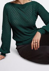 Femme assise portant un pull vert foncé texturé avec des motifs en losange et deux bagues à la main droite reposant sur sa jambe.