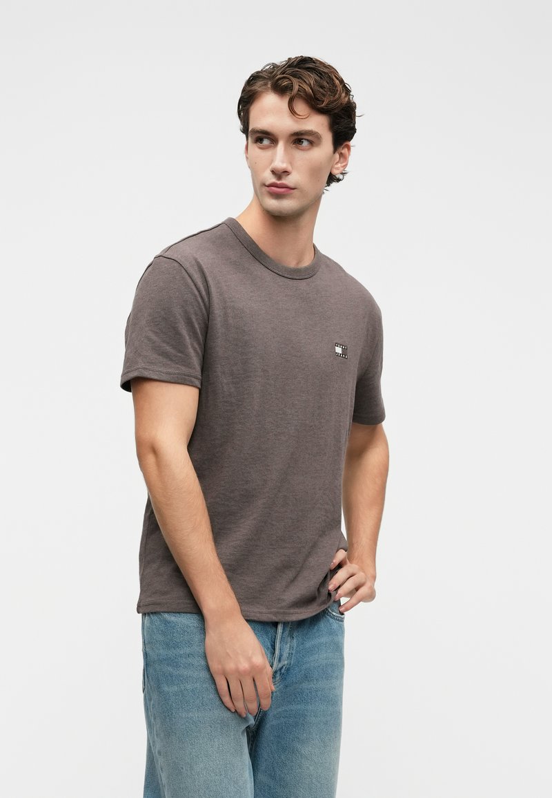 Ung mand med kort, krøllet hår iført en ensfarvet mørkegrå t-shirt og lyseblå jeans, der kigger til siden med hånden på hoften.