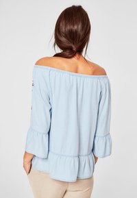 Blusa leggera azzurro chiaro con spalle scoperte, maniche a campana e orlo arricciato. Presenta un collo elasticizzato e dettagli ricamati sulle maniche.