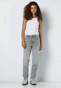 Jauna moteris su ilgais garbanotais plaukais, vilkinti baltais be rankovių marškiniais, šviesiai pilkais džinsais ir baltais sportiniais bateliais, stovi prieš paprastą baltą foną.