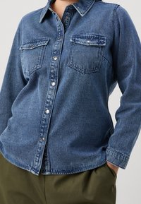Denim shirt in dark blue with long sleeves, a collared neckline, two chest pockets, and silver snap buttons. The fabric appears smooth and sturdy.