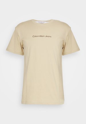Beżowy T-shirt z krótkim rękawem i okrągłym dekoltem z napisem „Calvin Klein Jeans” na środku klatki piersiowej.