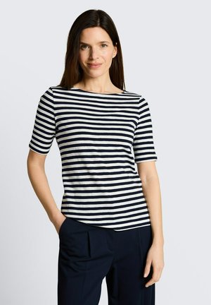 Kobieta o długich brązowych włosach, ubrana w czarno-białą paskowaną koszulkę z krótkim rękawem i ciemne spodnie, stoi z jedną ręką w kieszeni.