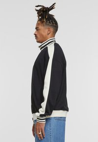 Sort sweatshirtjakke med hvide striber langs ærmerne, høj krave, sølvlynlås, ribbede manchetter og kant, båret over lyseblå jeans.