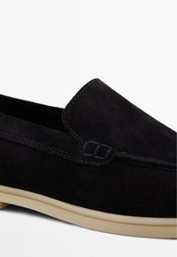 Scarpa slip-on in suede nera con punta arrotondata, suola in gomma di colore chiaro a contrasto e cuciture decorative lungo il bordo superiore.