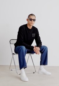 Pulover negru din tricot, cu logo-ul brodat "HUGO", asortat cu blugi albaștri și adidași albi. Modelul este așezat pe un scaun metalic.