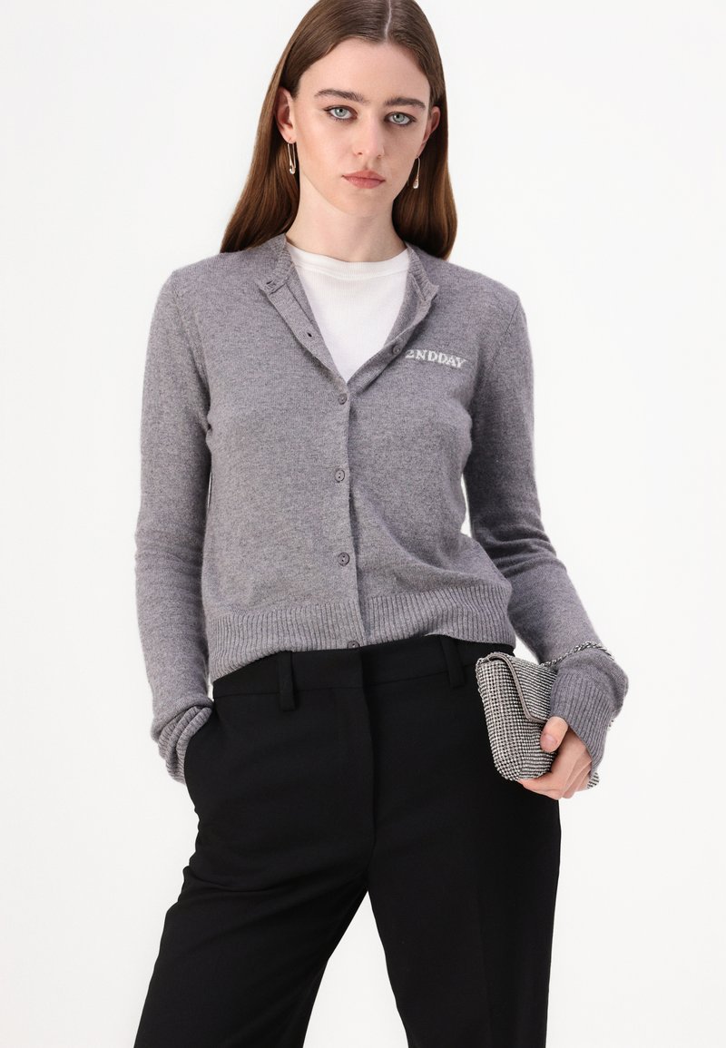 Grauer Wollcardigan mit V-Ausschnitt, vorderen Knöpfen und gerippten Bündchen. Mit kleinem "2NDDAY"-Stick auf der linken Brust. Schwarze taillierte Hose.