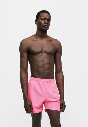 Homme torse nu avec une silhouette musclée portant un short de bain rose vif sur un fond blanc uni.