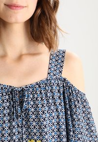 Niebieski i biały wzorzysty top typu off-shoulder z cienkimi ramiączkami, z detalem w postaci wiązania przy dekolcie i luźnym krojem.