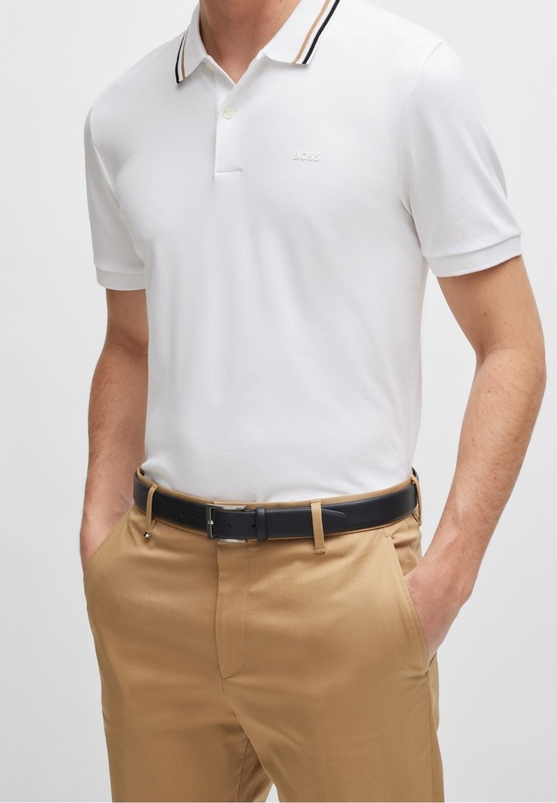 Polo blanc avec col rayé noir et beige, manches courtes et logo subtil. Associé à un pantalon marron clair et une ceinture noire.