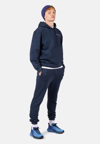 Felpa blu navy con tasca frontale e cordini, abbinata a pantaloni jogging blu navy e scarpe da ginnastica blu. Presenta calze bianche e un berretto blu.
