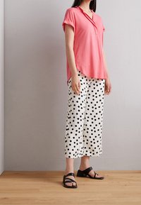 Helles rosa Kurzarmshirt mit V-Ausschnitt, kombiniert mit weißen weiten Hosen mit großen schwarzen Punktmustern; schwarze Sandalen mit Riemchen.