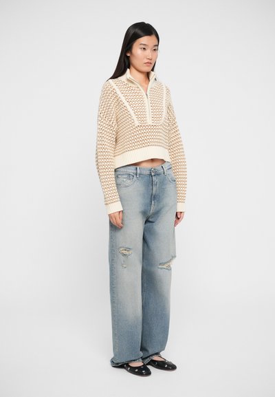 Beige knitted cropped sweater with a half-zip collar and textured pattern, paired with light blue distressed wide-leg jeans and black shoes.