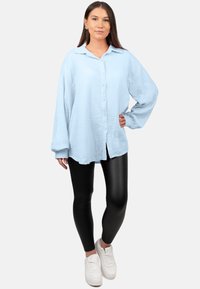Chemise boutonnée bleu clair en tissu texturé, coupe oversize et manches longues, associée à des leggings brillants noirs et des baskets blanches.