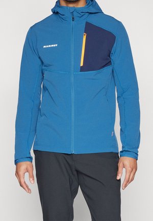 Mand iført blå hættetrøje med navy brystlomme og gul lynlås, kombineret med sorte bukser.