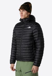 Chaqueta de plumas negra con baffles horizontales, cuello alto y cremallera en la parte delantera. Llevada con un gorro de camuflaje y pantalones verdes, que cuentan con bolsillos con cremallera.