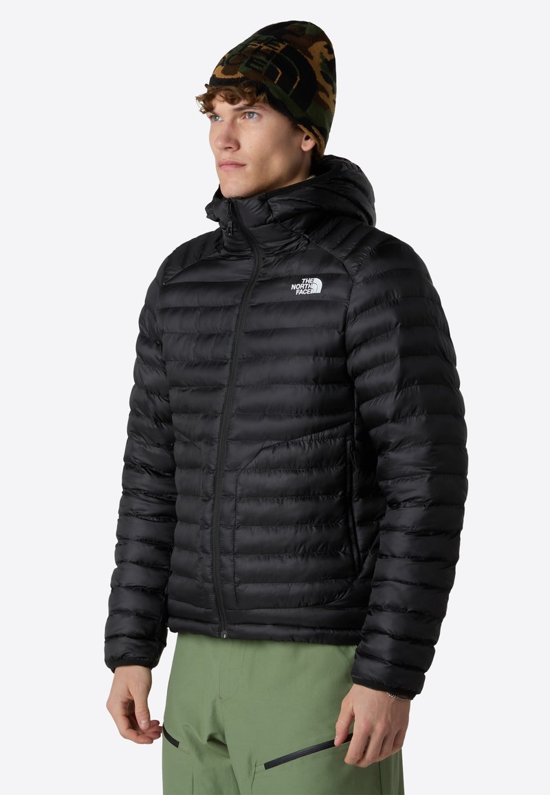 Chaqueta de plumas negra con baffles horizontales, cuello alto y cremallera en la parte delantera. Llevada con un gorro de camuflaje y pantalones verdes, que cuentan con bolsillos con cremallera.