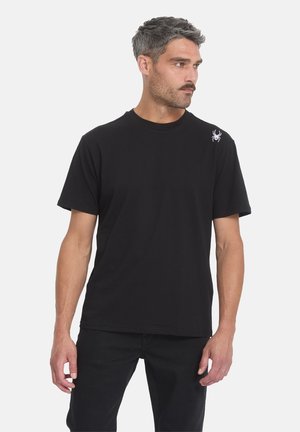 Homme aux cheveux gris courts et moustache, portant un t-shirt noir uni avec un petit logo d’araignée blanche sur l’épaule gauche, regardant vers la droite.