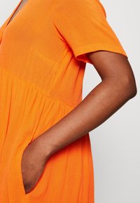 Orangenes Kurzarmkleid mit V-Ausschnitt, strukturiertem Stoff, geraffter Taille und seitlichen Taschen. Verfügt über eine Brusttasche.