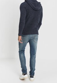 Marinblå stickad hoodie med en stor, vikbar krage. Matchad med ljusblå denimjeans och vita sneakers. Texturerad tygdetalj är synlig.