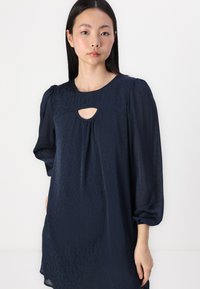 Blusa azul-marinho com detalhe de buraco de fechadura, mangas bufantes e uma silhueta fluida feita de um tecido leve e texturizado.